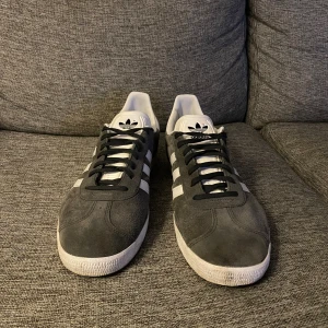 Adidas Gazelle grå sneakers - Adidas Gazelle sneakers i mörkgrå mocka med klassiska vita ränder på sidorna och vit häl. Platt sula med mönstrad undersida, svart snörning och rund tå. Ikonisk retrostil som funkar till streetwear och casual outfits. En liten defekt på hålen inte testat om det går bort! Jag tror det bara behöver en tvätt!