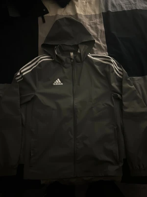 Grå Adidas vindjacka S - Snygg grå vindjacka från Adidas i storlek XS. Jackan har huva, hel dragkedja och klassiska vita ränder på axlarna. Adidas-logga på bröstet och två sidofickor. Perfekt för dig som gillar sportig stil och vill ha något lätt och funktionellt.