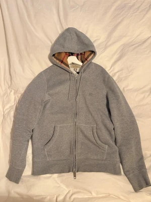 Burberry hoodie  - Säljer en grå hoodie från Burberry i storlek M. Tröjan har dragkedja, snörning och klassisk rutig insida på huvan. Tillverkad i mjuk bomull och polyester, med två fickor framtill och ribbade muddar. Perfekt för dig som gillar stilren design med lyxig touch.