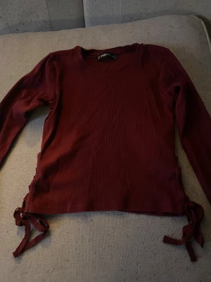 Vinröd ribbad tröja FB Sister XS - Snygg vinröd långärmad tröja från FB Sister i ribbad bomull. Tröjan har rund hals och coola snörningsdetaljer på båda sidorna vid midjan. Perfekt för dig som gillar en enkel men ändå unik look.