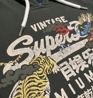 Superdry Hoodie - En riktigt fet Superdry hoodie med ett unikt och snyggt tryck, den är olivgrön ish med små detaljer som en liten patch på armen och liten text på snörerna. Den har inga defekter eller någon sorts skada. Kom pm för mått, inga skambud 