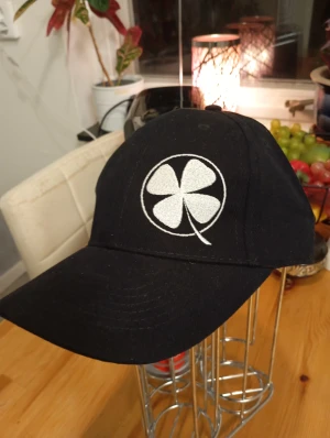 Ny keps - Svart keps med vit broderad klöver på framsidan och texten 'klovenas.se' på sidan. Klassisk böjd skärm och justerbar rem bak. Tillverkad i slitstark bomull, perfekt för dig som vill ha en clean och stilren look.