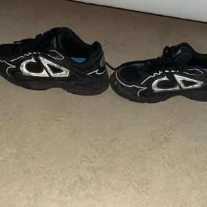 Svarta Dior sneakers med silverdetaljer tvättas inann frakt  - Säljer ett par svarta Dior sneakers med coola silvriga CD-loggor på sidorna och hälen. Skorna har snörning, platt sula och rund tå. Materialet är en mix av syntet och skinn, med mönstrade detaljer som ger en unik vibe. Perfekt för dig som vill sticka ut med designer på fötterna.