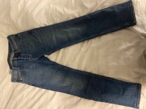 Blå jeans från Diesel straight fit - Säljer ett par klassiska blå jeans från Diesel med straight fit och normal passform. Jeansen har fem fickor, snygga slitningar och en tydlig Diesel-logga på bakfickan. Perfekt för dig som gillar tidlös stil och bekväma jeans.
