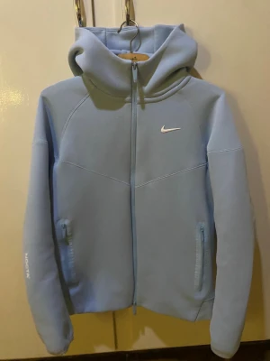Nike NOCTA ljusblå hoodie med dragkedja - Säljer en ljusblå hoodie från Nike NOCTA med hel dragkedja och huva. Tröjan har långa ärmar, två fickor med dragkedja och snygga vita logodetaljer på bröstet och ärmen. Perfekt för chill eller träning, riktigt clean design.