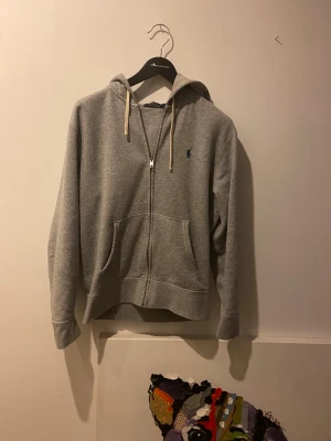 Grå hoodie från Polo Ralph Lauren - Snygg grå hoodie från Polo Ralph Lauren med dragkedja, huva och vita snören. Klassisk blå logga på bröstet och två stora fickor framtill. Perfekt för chill dagar och enkel att matcha med jeans eller joggers.
