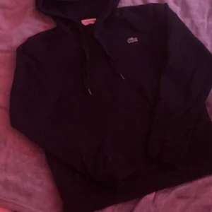 Svart hoodie från Lacoste med huva av - Svart hoodie från Lacoste med klassisk krokodillogga på bröstet. Tröjan har huva med snörning och långa ärmar. Perfekt för dig som gillar stilrena och bekväma plagg med sportig vibe.