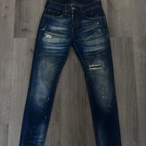 Dsquared2 byxor slim fit, storlek 44 - Säljer ett par Dsquared2 jeans slim i mörkblå tvätt med slitningar, lagade hål och coola färgstänk. Jeansen har klassisk femficksdesign, orange sömmar och smal passform. Något insydda i midjan. Perfekta för dig som gillar en streetig och avslappnad look.