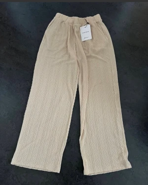 Beige mjuka byxor Vero Moda  - Snygga beige byxor från Vero Moda med bred resår i midjan och ett ribbat mönster. Byxorna har en relaxed passform och raka ben. Materialet är mjukt och ger en skön känsla. Nya med etikett. Sidofickor i fram. Benvidd ca 27cm. Midjemått ca 35cm. Innerbenslängd ca 77cm.