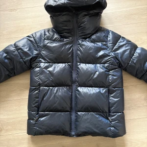 Blå pufferjacka från Everest - Säljer en svart pufferjacka från Everest med huva och markerad logga på ärmen. Jackan har en hög krage, quiltad design, dragkedja framtill och är tillverkad i glansig syntet. Perfekt för kalla dagar och har en clean, modern look.