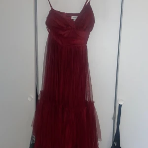 Vinröd långklänning med tyll från Nelly - Vinröd långklänning från Nelly med tunna axelband och vackert draperad tyll över hela klänningen. Klänningen har markerad midja, volangdetaljer nertill och dragkedja i ryggen. Perfekt för dig som vill sticka ut med en romantisk och elegant look.