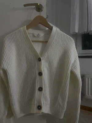 Vit ribbad kofta från H&M - Säljer en vit, ribbstickad kofta från H&M med stora svarta knappar framtill. Koftan har v-ringad hals och långa ärmar, perfekt för lager-på-lager. Materialet är mjuk stickad bomull som känns skönt mot huden.