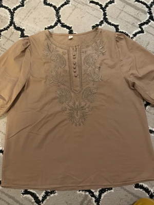 Beige broderad blus med puffärm - Säljer en beige blus från OZOC med broderade detaljer framtill och dekorativa knappar vid halsringningen. Blusen har puffiga trekvartsärmar och är tillverkad i ett tunt bomullstyg. Perfekt för dig som gillar boho-vibes och snygga detaljer.