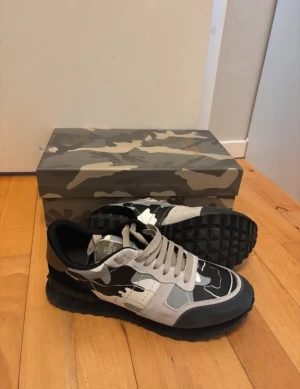 Valentino Garavani Rockrunner sneakers - Säljer ett par Valentino Garavani Rockrunner sneakers med grå, svart och vit camouflage-mönster. Skorna har platt sula med grovt mönster, rund tå och snörning. Materialmix av mocka och skinn ger en lyxig vibe. Box med matchande camo medföljer.