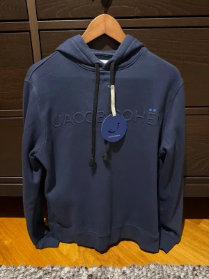 Mörkblå hoodie Jacob Cohën - Snygg mörkblå hoodie från Jacob Cohën med broderad logga på bröstet och svarta dragsnören. Klassisk huva och mjukt material, perfekt för en chill och stilren look. Diskreta detaljer och exklusiv känsla. Fint skick 