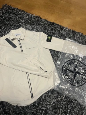  overshirt från Stone Island - Snygg beige overshirt från Stone Island med klassisk logotyp-patch på vänster ärm. Jackan har en rak passform, bröstficka med lock och svart dragkedja framtill. Materialet är lätt och syntetiskt, perfekt för lager-på-lager. Stilren och modern look.