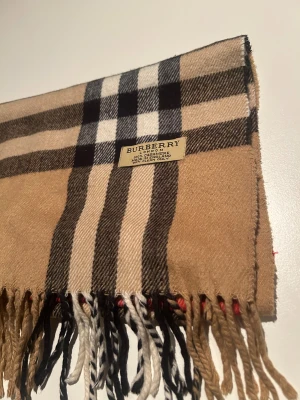 Rutig beige halsduk från Burberry - Klassisk halsduk från Burberry i 100% kashmir med det ikoniska rutiga mönstret i beige, svart, vitt och rött. Mjukt material och snygga fransar i ändarna. Perfekt accessoar för att lyfta din stil och hålla dig varm under kyliga dagar.