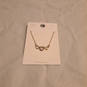 Guldfärgat halsband med hjärtan - Sött guldfärgat halsband från med tre hjärtan i mitten, där det mittersta hjärtat är dekorerat med gnistrande stenar. Kedjan är tunn och elegant, perfekt för att ge en romantisk touch till din outfit. Materialet är stål. Skriv till mig om du har frågor eller funderingar😉