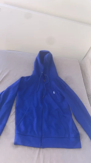 Blå hoodie från Polo Ralph Lauren - Snygg blå hoodie från Polo Ralph Lauren med klassisk vit broderad logga på bröstet. Tröjan har huva med snörning, dragkedja framtill och två fickor. Perfekt för dig som gillar stilren och sportig look.