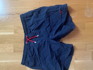 Mörkblå badshorts från GANT - Snygga mörkblå badshorts från GANT med röd snörning i midjan och diskret GANT-logga broderad på benet. Shortsen har elastisk midja och är perfekta för sommardagar vid stranden eller poolen. Säljer även några t shirts. Kom med pris