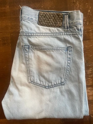 Ljusblå Our Legacy jeans med flätad patch - Snygga ljusblå jeans från Our Legacy med klassisk femficksdesign och en unik flätad patch i mocka på bakfickan. Jeansen har raka ben och normal passform, tillverkade i mjukt denimtyg. Perfekt för dig som gillar stilrena detaljer och ljusa toner.