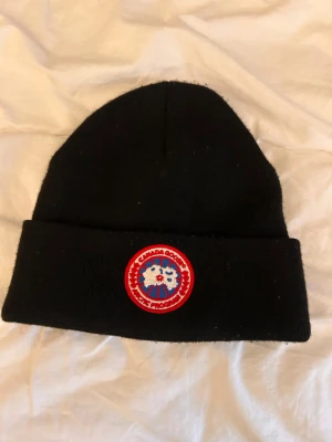 Svart mössa f - Svart ribbstickad mössa med klassisk rund logotyp-patch framtill i rött, vitt och blått. Tillverkad i 100% bomull, perfekt för kalla dagar och snygg till streetwear-looken