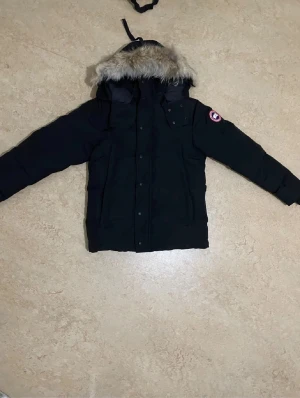 Svart dunjacka från Canada Goose S - Säljer en svart Canada Goose dunjacka i storlek S med avtagbar huva och fuskpäls. Jackan har klassisk patch på ärmen, stora fickor och stängs med både dragkedja och knappar. Perfekt för kalla vinterdagar och riktigt snygg streetstil.