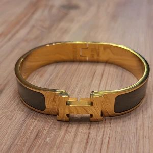 Armband med H-detalj, clic H  - Snyggt och ikoniskt armband i guldtonad metall med grön-gråa emaljpaneler. Armbandet har den klassiska H-formade låsdelen framtill och en stilren, rundad form som sitter bekvämt runt handleden. Perfekt för dig som gillar lyxiga accessoarer med statement. 6 cm i diameter, 5 cm på höjden. 