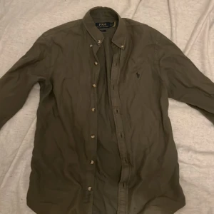 Olivgrön skjorta från Polo Ralph Lauren - Snygg olivgrön skjorta från Polo Ralph Lauren med klassisk button down-krage och broderad logga på bröstet. Skjortan har långa ärmar och knäppning framtill. Perfekt för en stilren och avslappnad look.
