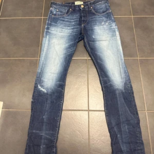 Blå jeans från Scotch & Soda - Säljer ett par snygga blå jeans från Scotch & Soda med coola slitningar och tvättade detaljer. Modellen har klassisk femficksdesign, raka ben och normal passform. Jeansen har en läderpatch med logga bak i midjan och är tillverkade i mjukt denimtyg.