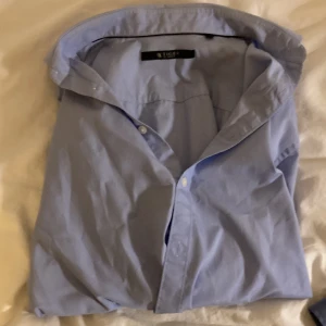 Ljusblå skjorta från Tiger of Sweden - Klassisk ljusblå skjorta från Tiger of Sweden i bomull. Skjortan har lång ärm, knappar framtill och en stilren krage. Perfekt för dig som gillar en clean och tidlös look.