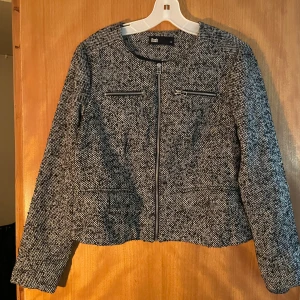 Grå rutig kavajjacka från Flash - Snygg grå kavajjacka från Flash med rutigt mönster och dragkedja framtill. Jackan har två dekorativa dragkedjor vid bröstet och är långärmad med en croppad passform. Perfekt för dig som gillar stilrena detaljer och vill ha en jacka som sticker ut.