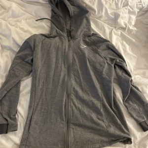 Grå hoodie från Gymshark L - Säljer en grå hoodie från Gymshark i storlek L. Hoodien har hel dragkedja, huva med snörning och lång ärm. Materialet är mjukt och lätt, perfekt för träning eller chill. Diskret Gymshark-logga broderad på bröstet. Enkel och stilren design.