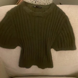 Olivgrön stickad tröja från Zara - Supermjuk olivgrön stickad tröja från Zara med korta ärmar och ribbat mönster. Tröjan har en lite fluffig känsla och sitter snyggt figurnära i midjan. 