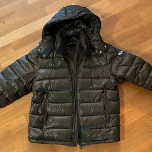 Mörkgrön pufferjacka från Bvaldi - Snygg mörkgrön pufferjacka från Bvaldi med huva och coolt BB-emblem på ärmen. Jackan har två dragkedjeförsedda fickor, quiltad design och en riktigt skön, vadderad känsla. Perfekt för kalla dagar när du vill hålla stilen. Jackan är som ny!