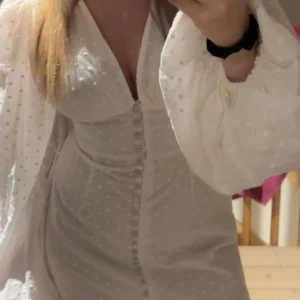 Vit prickig kortklänning från Adoore - Supersöt vit kortklänning från Adoore med diskret prickigt mönster, v-ringad hals och långa puffärmar. Klänningen har knappar framtill och är figurnära med lätt volym i ärmarna. Perfekt för dig som gillar romantisk och trendig stil. Super fin till student osv. Har en knapp som lossnat men skickar med den!