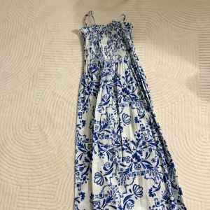 Vit och blå långklänning med blommor - Säljer en superfin långklänning med smala axelband och smock upptill. Klänningen är vit med blått blommigt mönster och har en luftig, flowy passform. Perfekt för varma dagar och har en riktigt fräsch vibe. Materialet känns lätt och bekvämt mot huden.