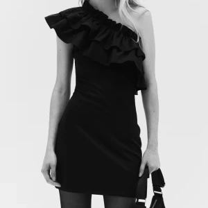 Svart oneshoulder klänning med volang - Snygg svart kortklänning med oneshoulder-design och stora volanger längs axeln. Klänningen har en tight passform och är perfekt för dig som vill sticka ut med en cool och trendig look. Materialet är mjukt och följsamt, vilket gör den bekväm att bära hela kvällen. Helt slutsåld, aldrig använd, bara testad. 