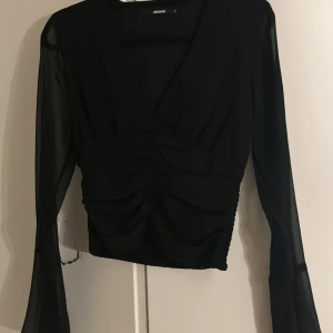 Svart blus med v-ringning från Bik Bok - Säljer en svart blus från Bik Bok med v-ringad front och snygg rynkning över magen. Blusen har långa, transparenta ärmar och är croppad i modellen. Perfekt för dig som gillar stilrena och trendiga plagg med lite extra edge.