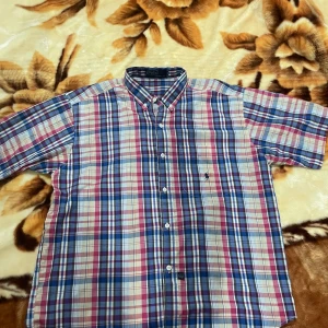 Rutig kortärmad skjorta från Polo XL - Säljer en färgstark rutig skjorta från Polo i storlek XL. Skjortan har korta ärmar, klassisk button-down krage och knappar framtill. Materialet är 100% bomull och färgerna går i blått, rött, vitt och inslag av beige. Polo-loggan är broderad på bröstet.