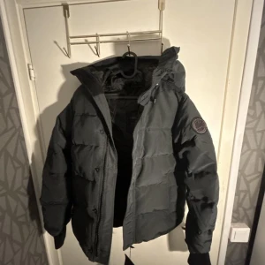 Svart pufferjacka från Canada Goose - Säljer en svart pufferjacka från Canada Goose med huva och snyggt märke på ärmen. Jackan har dragkedja och knappar framtill, quiltad design och är perfekt för kalla dagar. Insidan har äkta etikett och detaljerad märkning. 