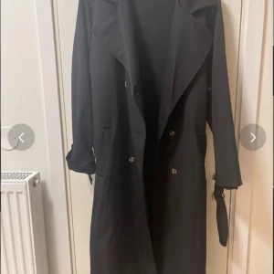 Svart trenchcoat med bälte - Snygg svart trenchcoat med klassisk krage, knappar framtill och bälte i midjan. Ärmarna har justerbara spännen och jackan har fickor på sidorna. Perfekt för dig som gillar stilrena och tidlösa plagg med coola detaljer.