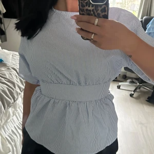 Blårandig omlottblus med knyt - Säljer en blårandig omlottblus med korta ärmar och knyt i midjan. Blusen har en luftig passform och är tillverkad i mjuk bomull. Snygg v-ringning framtill och volangdetaljer vid midjan ger en chill och trendig vibe.