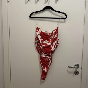 Vit och röd blommig body H&M - Säljer en snygg body från H&M med tunna axelband och draperad front. Bodyn är vit med stora röda blommor och har en figurnära passform. Perfekt för dig som gillar färgstarka mönster och vill sticka ut lite extra.