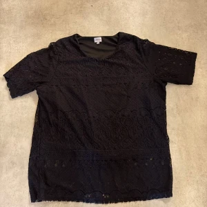 Svart spetsblus från Amalia XL - Svart blus från Amalia i storlek XL, tillverkad i spets med snygga broderade detaljer och korta ärmar. Blusen har rund halsringning och ett genomgående mönster i tyget som ger en cool och elegant vibe.
