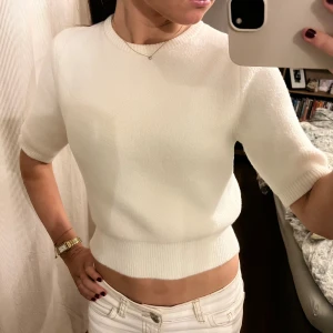 Vit stickad kortärmad tröja - Supersnygg vit stickad tröja med korta ärmar och rund hals. Modellen är croppad och har ribbad kant vid midjan och ärmslut. Perfekt för dig som gillar en clean och fräsch look med lite edge.