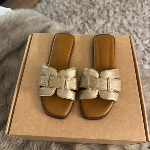 Guldiga sandaler med fyrkantig tå - Snygga sandaler i guldigt skinn med fyrkantig tå och bred rem med flätad detalj. Platt sula och öppen design, perfekta för varma dagar. Stilsäkra och trendiga med en exklusiv känsla🩷    Obs, box tillkommer ej