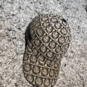 Salvatore Ferragamo beige keps - Snygg beige keps från Salvatore Ferragamo med mörkgrått, grafiskt mönster över hela kepsen och svart läderdetalj baktill med logotyp. Kepsen har böjd skärm och justerbar passform. Perfekt för dig som gillar designeraccessoarer med unik look.