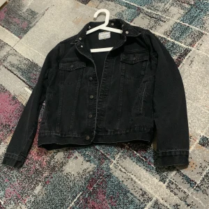 Svart jeansjacka från SikSilk - Svart jeansjacka från SikSilk i klassisk modell med knappar framtill, två bröstfickor med lock och sidofickor. Jackan har en snygg krage och detaljerad sömnad, samt en brun läderpatch bak vid nederkanten. Perfekt för dig som gillar streetstyle.