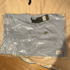 Ljusblå t-shirt från Lyle & Scott - Snygg ljusblå t-shirt från Lyle & Scott med klassisk gul logga på bröstet. T-shirten har rund hals och är kortärmad, perfekt för en clean och stilren look. Tillverkad i mjuk bomull för maximal komfort.